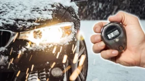 A person holding a stopwatch while a black car with snow on top of it warms up in the snow